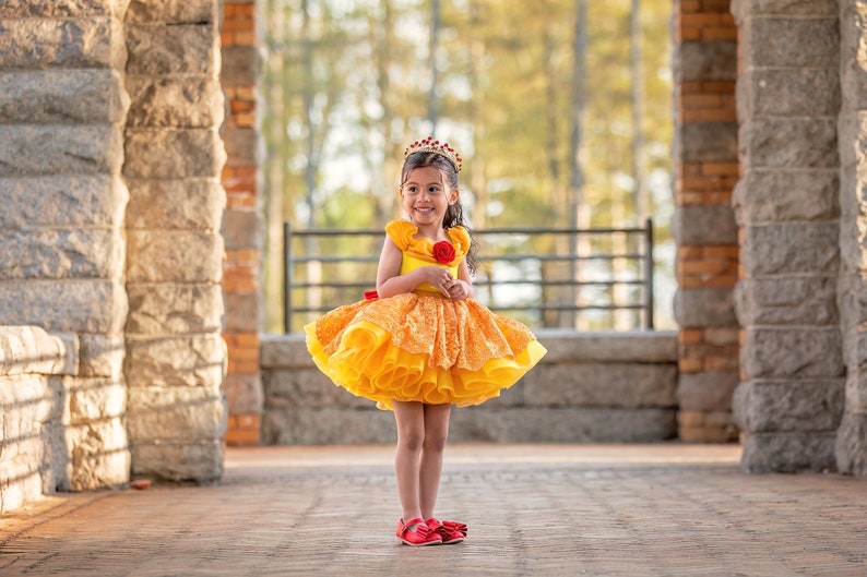May include: A young girl wearing a yellow and orange dress with a red rose and a crown. The dress has a layered skirt and ruffled sleeves. She is wearing red shoes and standing in front of a stone archway.