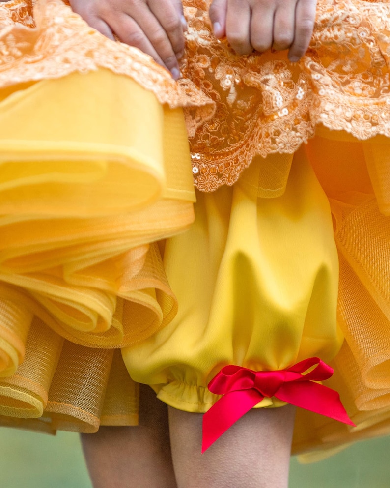 May include: A close-up of a yellow dress with a layered skirt and a red bow. The dress has a lace and sequin bodice. The lower part of the dress is a bright yellow color.