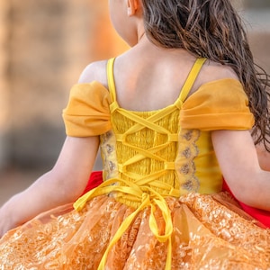 May include: A yellow and orange dress with a lace overlay and a corset-style back. The dress features off-the-shoulder sleeves and a ribbon tie. The dress is designed for a child.