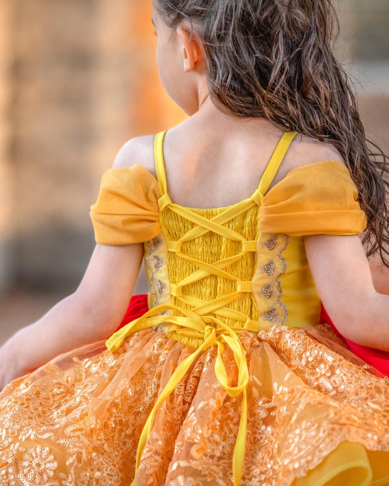 May include: A yellow and orange dress with a lace overlay and a corset-style back. The dress features off-the-shoulder sleeves and a ribbon tie. The dress is designed for a child.