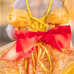 May include: A yellow and orange dress with a large red bow on the back. The dress features a lace-trimmed skirt and puff sleeves. The back of the dress has a corset-style design.