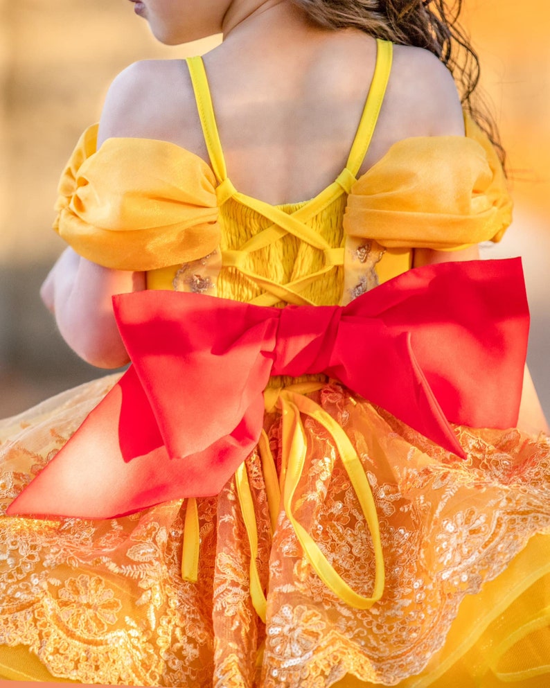 May include: A yellow and orange dress with a large red bow on the back. The dress features a lace-trimmed skirt and puff sleeves. The back of the dress has a corset-style design.