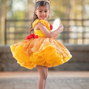 May include: A young girl in a yellow and orange princess dress with a red bow and matching red shoes. She wears a gold crown with red jewels and holds a small mirror. The dress has a layered skirt and a red rose detail.