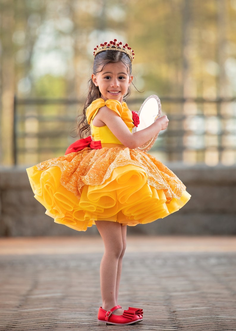 May include: A young girl in a yellow and orange princess dress with a red bow and matching red shoes. She wears a gold crown with red jewels and holds a small mirror. The dress has a layered skirt and a red rose detail.