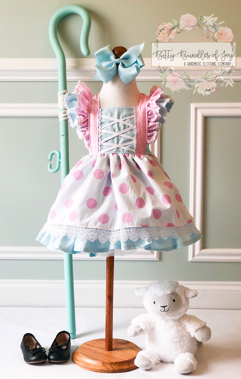 May include: A white dress with pink polka dots and blue accents. The dress has a lace trim and a pink and blue bodice with a criss-cross design. The dress is on a wooden stand with a shepherd's crook and a white plush lamb toy.