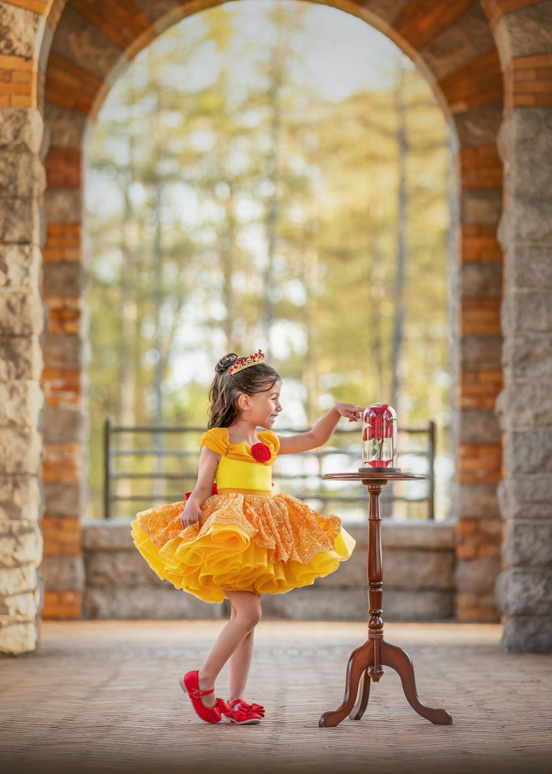 May include: A young girl in a yellow and orange dress with a red rose detail and a gold crown. She is standing near a small wooden table with a glass dome containing a red rose. The dress has a layered skirt and short sleeves. The girl is wearing red shoes.