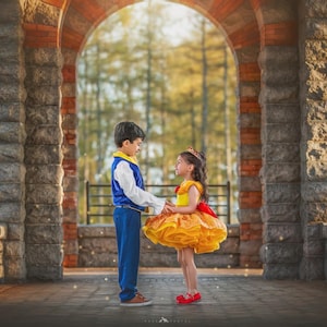 May include: Two children dressed in fairytale costumes stand facing each other under an arched stone structure. The boy wears a blue and white outfit, while the girl is in a yellow dress with a red bow. The background features a blurred view of trees and sunlight.