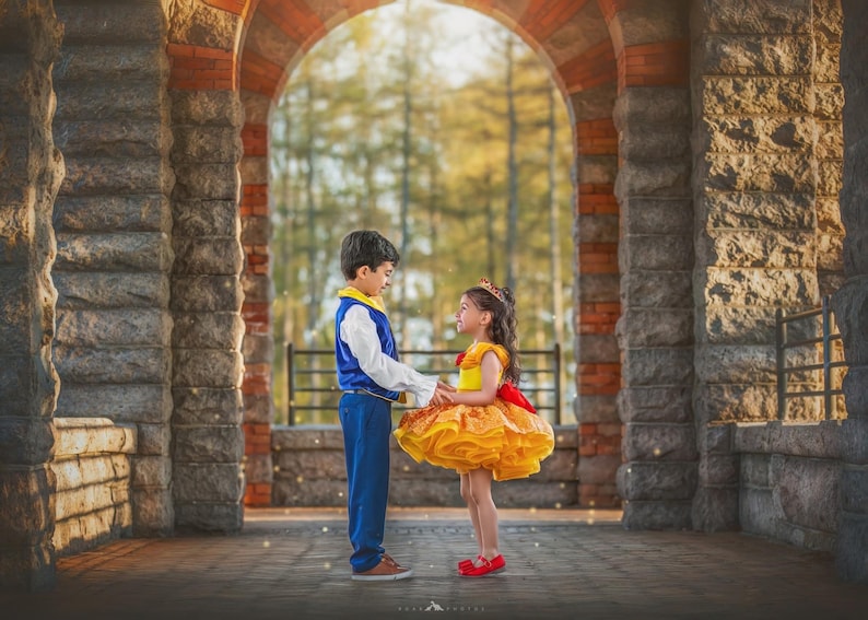 May include: Two children dressed in fairytale costumes stand facing each other under an arched stone structure. The boy wears a blue and white outfit, while the girl is in a yellow dress with a red bow. The background features a blurred view of trees and sunlight.