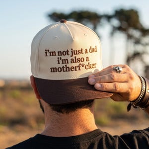 May include: A beige and brown baseball cap with the text "I'm not just a dad I'm also a motherf*cker" embroidered in brown. The cap has a curved brim and a button on top.