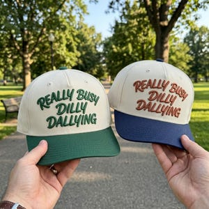 May include: Two baseball caps with off-white crowns and embroidered text reading "REALLY BUSY DILLY DALLYING". One cap features a dark green brim and matching green embroidery, while the other has a navy blue brim and brown embroidery.