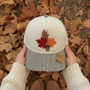 May include: A cream-colored baseball cap featuring an embroidered design of a brown acorn and three autumn leaves in shades of red, orange, and brown. The brim of the cap has a vertical black and white pinstripe pattern.