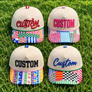 May include: Four beige baseball caps with structured crowns and colorful patterned brims. Each cap features the word CUSTOM embroidered on the front in different fonts and colors, including red, pink, black, and blue, with matching top buttons.