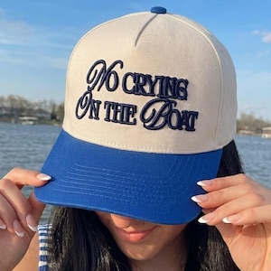 May include: A baseball cap with a cream-colored crown and a blue brim. The cap features the embroidered phrase "No Crying On The Boat" in navy blue script. The cap has a blue button on top.