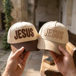 May include: Two baseball caps with the word "JESUS" embroidered on the front. The caps have a beige crown and a brown brim. One cap has brown lettering, and the other has beige lettering. The caps are held up in front of a blurred background.