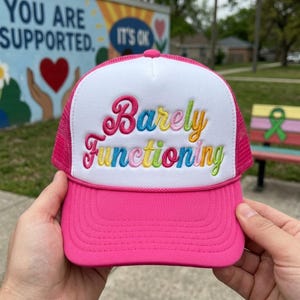 May include: A bright pink trucker hat with a white foam front panel. The text Barely Functioning is embroidered in a multi-colored script font across the center. The hat features a pink mesh back, a pink brim with stitching, and a pink button on top.