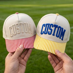 May include: Two baseball caps with beige crowns and the word CUSTOM embroidered on the front. The left cap features a red and white striped brim, while the right cap has a yellow and white striped brim. Both caps have a small button on top.