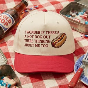 May include: A baseball cap with a cream-colored crown and a red brim. The cap features the text "I WONDER IF THERE'S A HOT DOG OUT THERE THINKING ABOUT ME TOO" and an illustration of a hot dog.