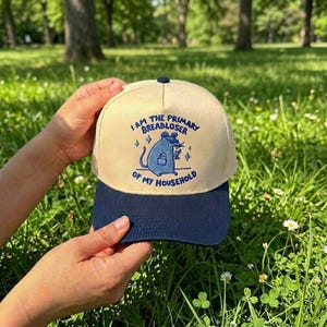 May include: A beige and navy blue baseball cap with the text "I AM THE PRIMARY BREADLOSER OF MY HOUSEHOLD" and a cartoon rat illustration. The cap has a curved brim and a button on top.