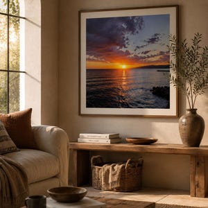 Può includere: Una stampa incorniciata di un tramonto sull'oceano, con vibranti tonalità arancioni e rosse nel cielo. L'opera d'arte è appesa sopra una panca di legno con un cesto intrecciato e oggetti decorativi. Un vaso con vegetazione è a destra.