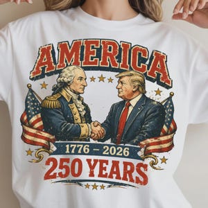 Puede incluir: Camiseta blanca con un dise&ntilde;o gr&aacute;fico. El dise&ntilde;o presenta la palabra "AMERICA" en rojo, con letras desgastadas. El gr&aacute;fico incluye ilustraciones de George Washington y Donald Trump d&aacute;ndose la mano, con banderas americanas y el texto "1776-2026 250 YEARS".