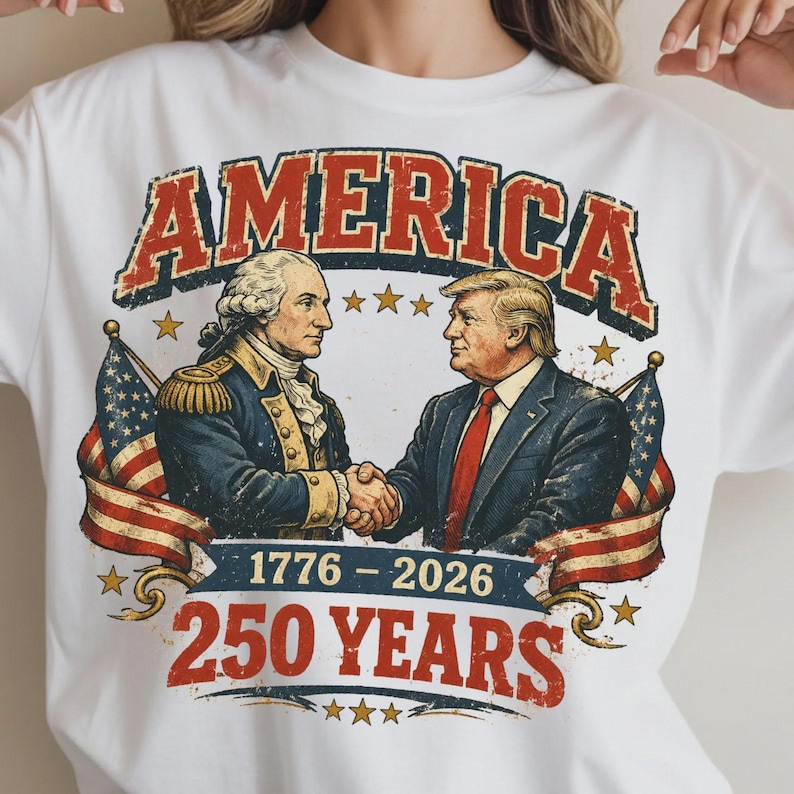 Puede incluir: Camiseta blanca con un dise&ntilde;o gr&aacute;fico. El dise&ntilde;o presenta la palabra "AMERICA" en rojo, con letras desgastadas. El gr&aacute;fico incluye ilustraciones de George Washington y Donald Trump d&aacute;ndose la mano, con banderas americanas y el texto "1776-2026 250 YEARS".