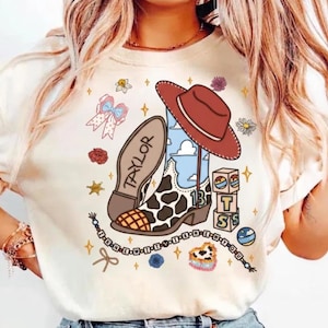 Pode incluir: Uma t-shirt cor de creme com uma impress&atilde;o gr&aacute;fica de uma bota de cowboy com manchas de vaca, um chap&eacute;u de cowboy vermelho, um la&ccedil;o, blocos de letras e um colar de contas. A bota exibe o nome TAYLOR e o n&uacute;mero 13 com nuvens.