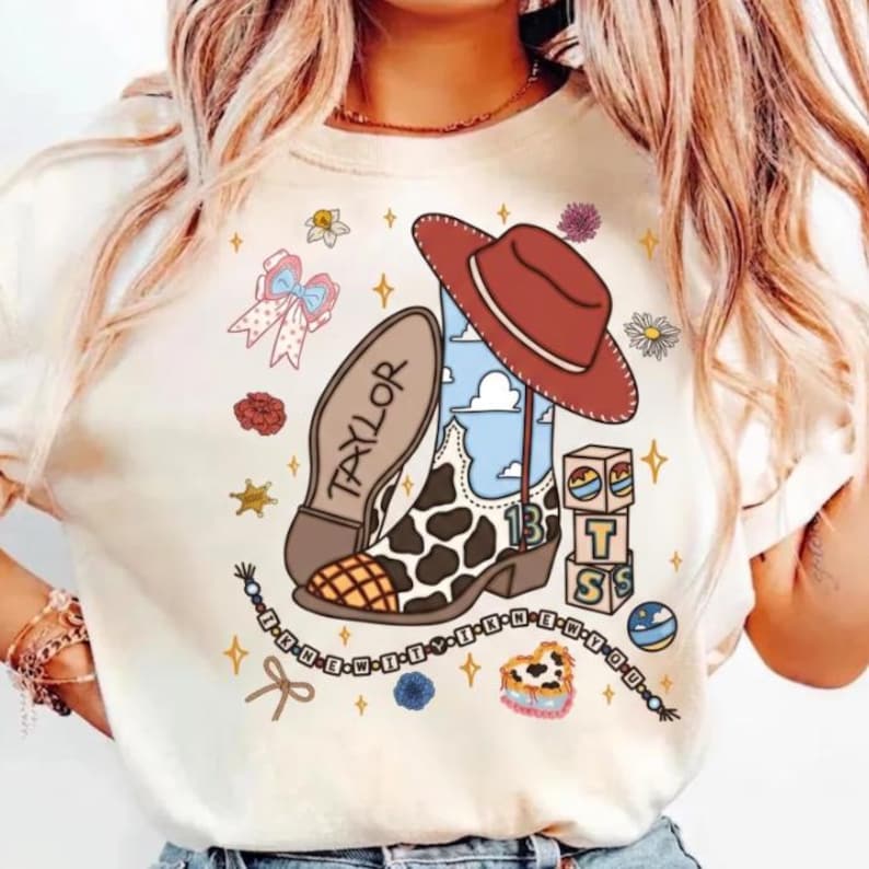 Pode incluir: Uma t-shirt cor de creme com uma impress&atilde;o gr&aacute;fica de uma bota de cowboy com manchas de vaca, um chap&eacute;u de cowboy vermelho, um la&ccedil;o, blocos de letras e um colar de contas. A bota exibe o nome TAYLOR e o n&uacute;mero 13 com nuvens.