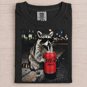 May include: A black crew neck t-shirt featuring a graphic print of a raccoon wearing black sunglasses and drinking from a red Coca-Cola Zero can with a white straw. The shirt has a Comfort Colors brand label printed on the inside of the neck collar.
