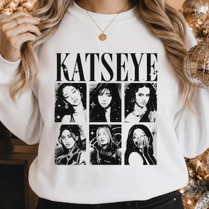 May include: A white crewneck sweatshirt featuring the black text KATSEYE at the top, followed by a grid of six black and white illustrated portraits of individuals, each framed within a square, accented with small star and circular graphic details.