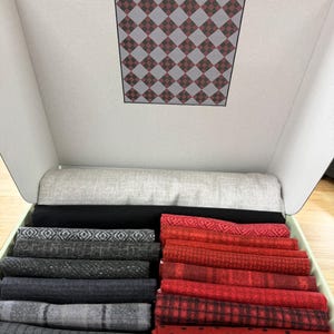 May include: A white cardboard box containing folded fabric pieces. The lid features a diamond pattern in red and gray. Inside, a light gray fabric roll sits above two columns of folded textiles: one column in shades of black and gray, and the other in various red patterns including plaid, dots, and geometric designs.