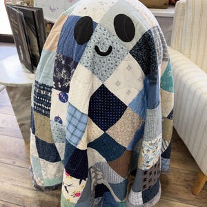 Pode incluir: Uma colcha em forma de fantasma feita de vários quadrados de tecido azul, branco e bege. A colcha apresenta dois olhos ovais pretos e uma boca curva preta. Vários retalhos de tecido exibem padrões, incluindo estampas florais, xadrez e logotipos da NASA.