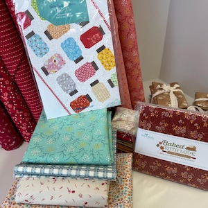 May include: A collection of quilting fabric bundles and a pattern booklet titled Yes You Can Table Runner Pattern by Lori Holt. The fabrics feature various floral, gingham, and geometric prints in shades of red, teal, yellow, and cream. A square fabric stack is labeled Baked With Love by Riley Blake.