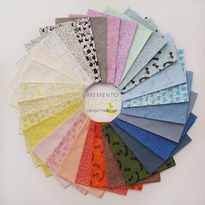 Op de afbeelding: Een cirkelvormige waaier van rechthoekige stoffenstalen met diverse patronen en kleuren, waaronder geel, oranje, groen, blauw en neutrale tinten. Een centraal wit rond label toont de tekst MEMENTO en carolyn friedlander.