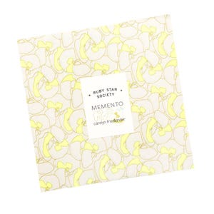 May include: A square stack of fabric squares featuring a repeating pattern of light grey outlines and pale yellow shapes. A white label in the centre displays the text Ruby Star Society, Memento, and Carolyn Friedlander in black lettering.
