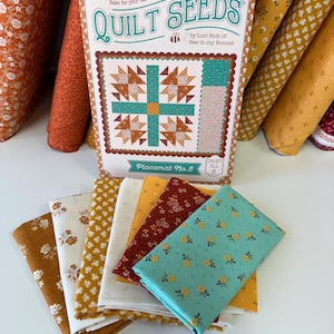 May include: A Quilt Seeds Placemat No. 8 pattern card by Lori Holt of Bee in my Bonnet, featuring a quilt illustration and text. Below the card are several folded fabric swatches in shades of mustard yellow, white, dark red, and teal with floral prints.