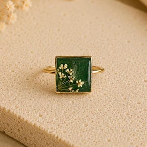 May include: A gold-toned ring featuring a square-shaped bezel with a deep green background. Encased within the green resin is a cluster of small, delicate white pressed flowers and fine, wispy botanical filaments arranged in a fan-like pattern.