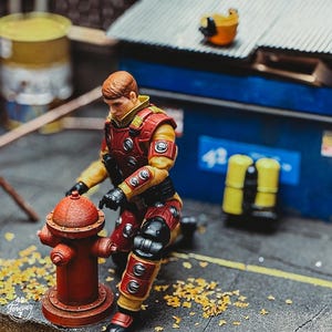 May include: A detailed action figure of a character in a red and yellow uniform kneels beside a red fire hydrant. The figure has black gloves and knee pads. The background includes a blue dumpster and a yellow barrel.