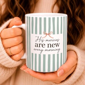 May include: A white ceramic mug featuring vertical sage green stripes and a central graphic of a small pink bow. The text "His mercies are new every morning" is printed in black script and serif lettering on the front of the mug.