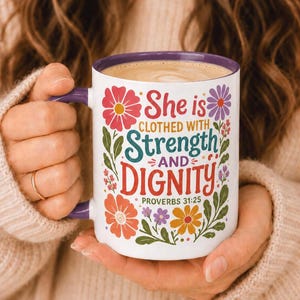 May include: A white ceramic mug with a purple handle and rim, featuring a colorful floral design and the text "She is CLOTHED WITH Strength AND DIGNITY PROVERBS 31:25" in various fonts and colors including red, teal, and orange on the front surface.