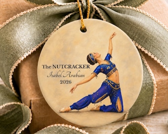Könnte beinhalten: Ein rundes Keramikornament mit einer Tänzerin in einem blauen Kostüm im arabischen Stil, die auf einem Bein kniet. Der Text lautet The NUTCRACKER, Isabel Arabian und 2026. Ein goldener Faden ist oben befestigt, umgeben von einem breiten olivgrünen Band.
