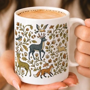 May include: A white ceramic mug filled with coffee, featuring a detailed illustration of a dark blue deer, a green deer, a brown fox, a rabbit, and various birds nestled among intricate green and brown leafy floral vines and small flowers.