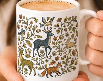 May include: A white ceramic mug filled with coffee, featuring a detailed illustration of a dark blue deer, a green deer, a brown fox, a rabbit, and various birds nestled among intricate green and brown leafy floral vines and small flowers.