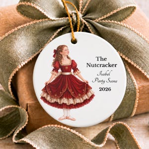 May include: A round white ceramic ornament featuring a girl in a red dress with gold trim, text reading The Nutcracker, Isabel Party Scene, and 2026, suspended by a gold string against a background of a brown gift with a green ribbon.