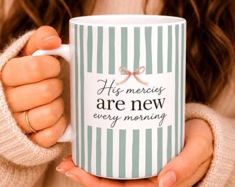 May include: A white ceramic mug featuring vertical sage green stripes and a central white panel. The text "His mercies are new every morning" is printed in black script and serif fonts, accented by a small, light pink bow illustration above the text.