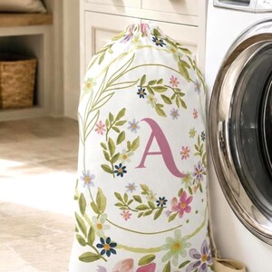 May include: A white fabric laundry bag with a drawstring closure, featuring a printed pattern of colorful watercolor-style flowers and green leaves arranged in a circular wreath design, with a large, centered, serif-style letter A in pink.