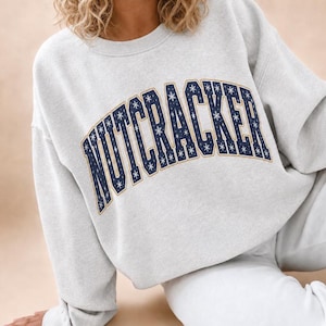 May include: A light grey crewneck sweatshirt featuring the word NUTCRACKER in a dark blue collegiate-style font. The letters are filled with a white snowflake pattern and outlined in a thin gold border, creating a festive winter aesthetic.