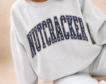 Könnte beinhalten: Ein hellgraues Sweatshirt mit Rundhalsausschnitt und dem Wort NUTCRACKER in einer dunkelblauen Schriftart im College-Stil. Die Buchstaben sind mit einem weißen Schneeflockenmuster gefüllt und mit einem dünnen goldenen Rand versehen.