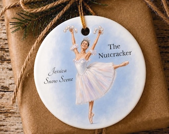 Könnte beinhalten: Ein rundes, weißes Keramikornament mit einer gemalten Ballerina im weißen Tutu und erhobenen Armen. Der Text lautet The Nutcracker und Jessica Snow Scene. Das Ornament hängt an einer goldenen Kordel vor einem braunen, in Papier gewickelten Hintergrund.
