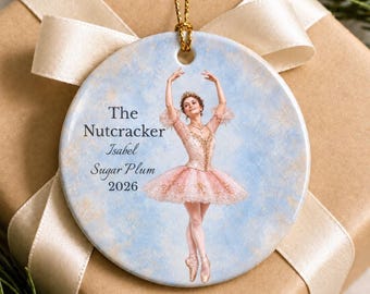 May include: A round ceramic ornament featuring a ballerina in a pink tutu against a light blue background. The text reads The Nutcracker, Isabel, Sugar Plum, 2026. A gold string is attached to the top for hanging, and the ornament is white.