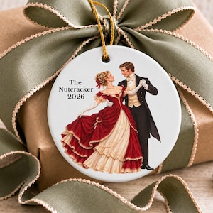 May include: A round white ceramic ornament featuring an illustration of a couple dancing. The text reads The Nutcracker 2026. The person on the left wears a red and cream gown, and the person on the right wears a black tuxedo with a gold vest.